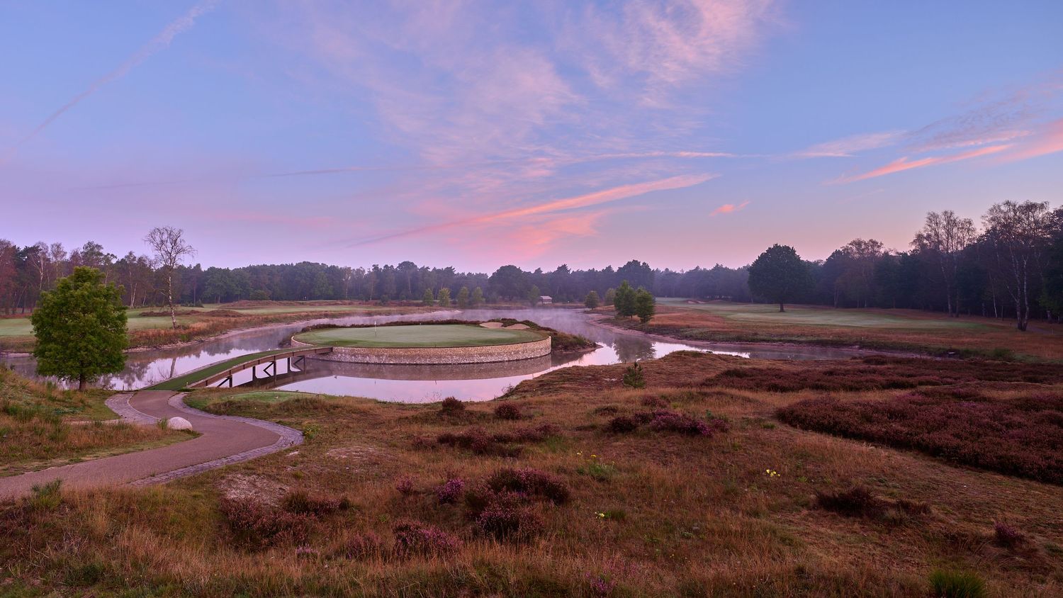 Aerial sunset view of a winding golf course with a pond, heathland, and colorful pink sky.