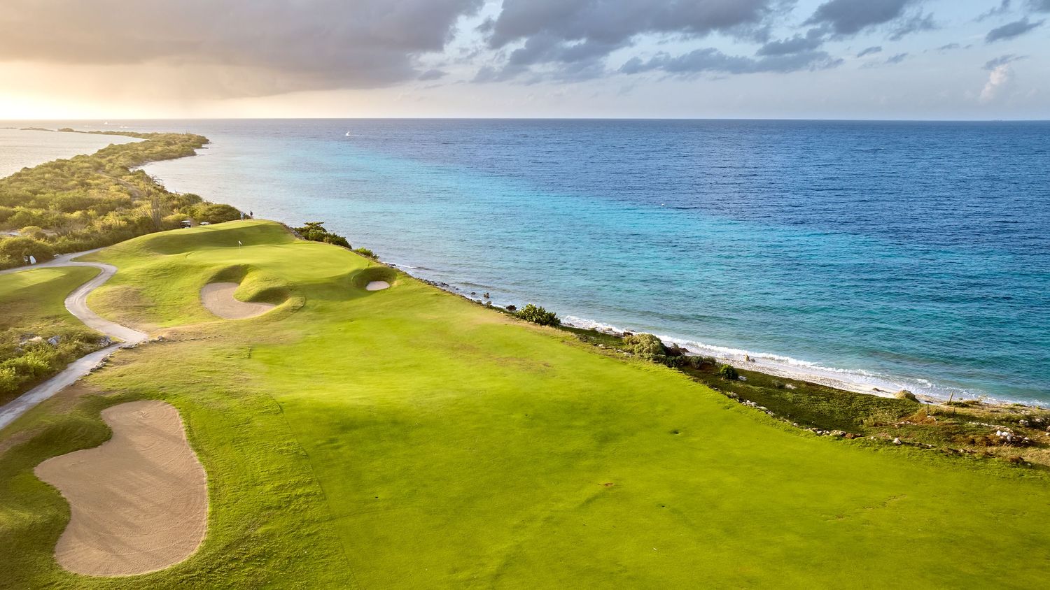 Aerial view of a lush green golf course along a stunning turquoise Caribbean coastline at sunset.