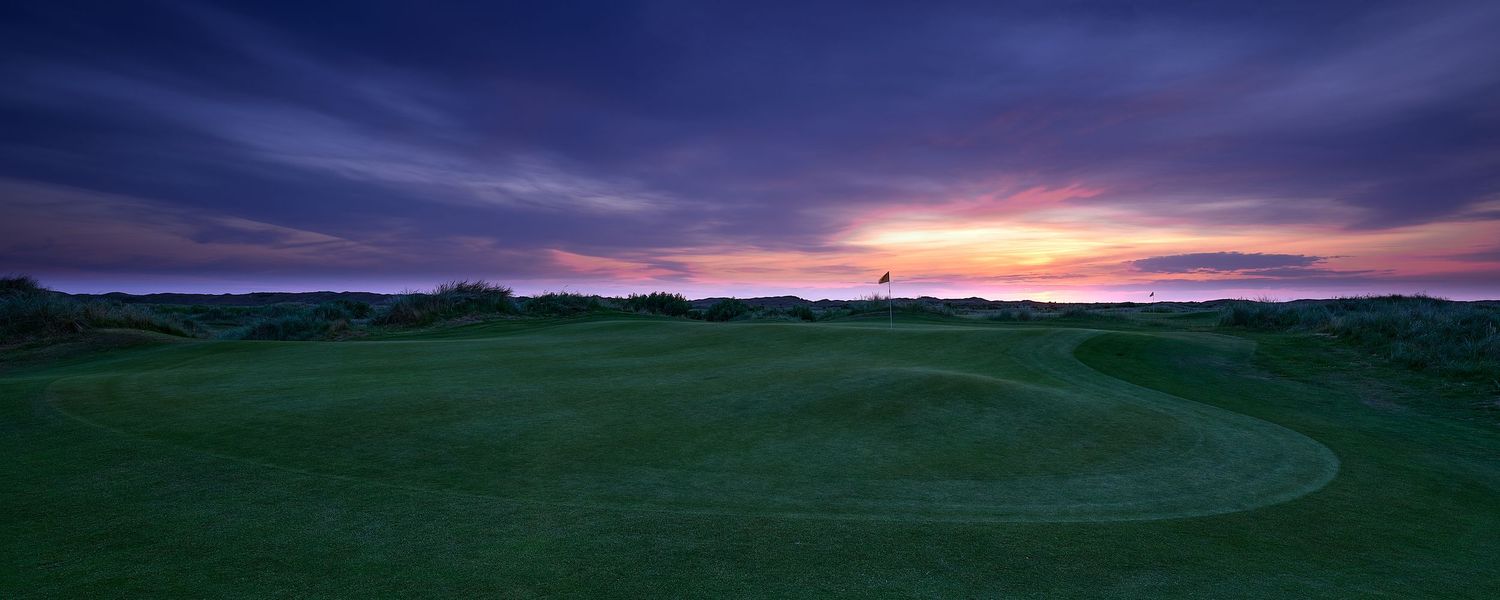 Golf course green at dramatic purple sunset with flag pin visible against colorful sky over coastal links course.