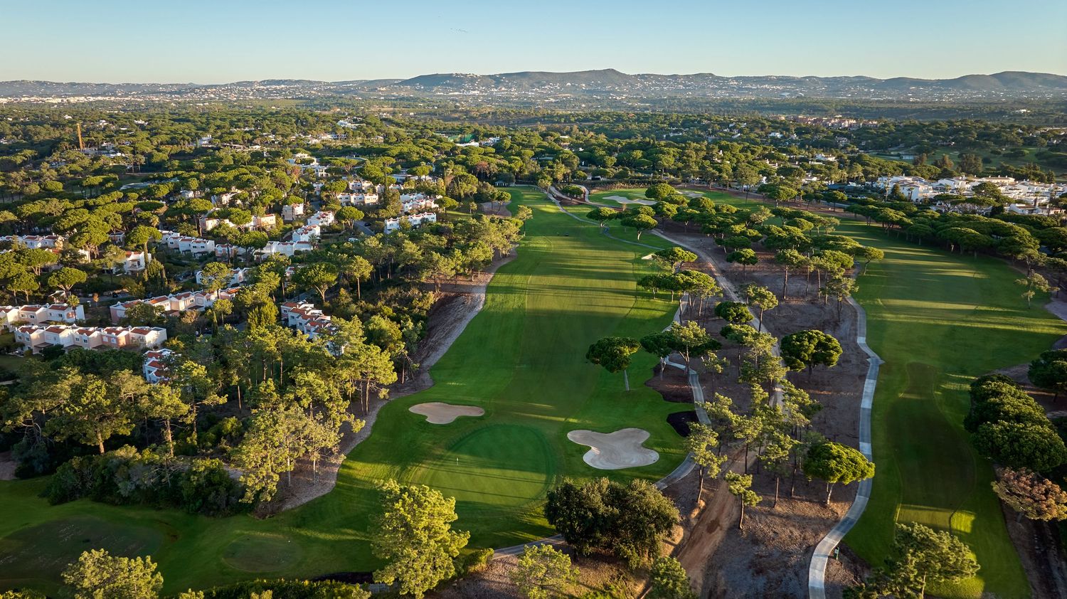 Aerial view of a lush golf course surrounded by green trees and residential areas with mountains in the background.