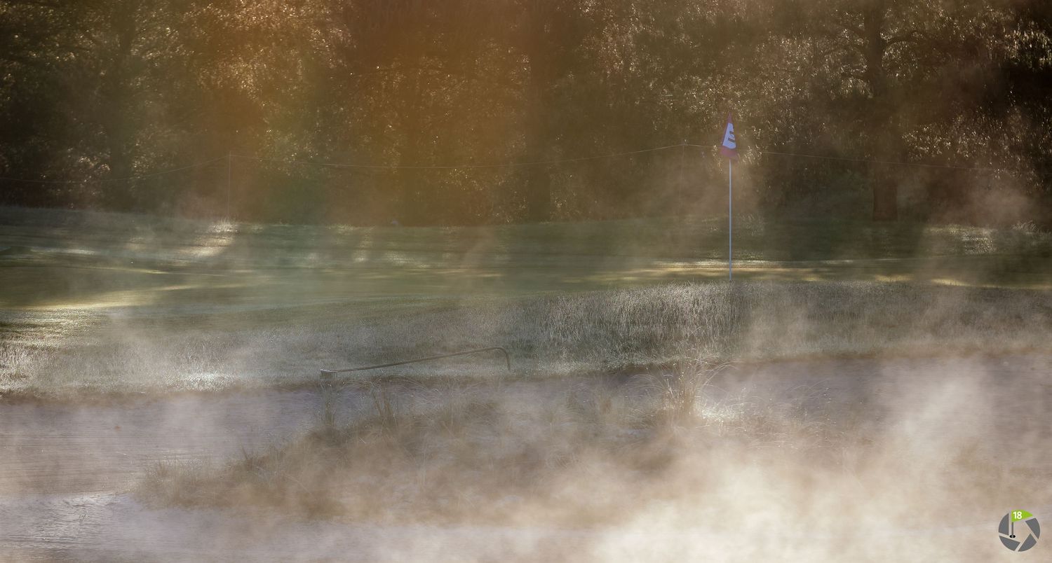Misty golf course green with a blue flag pin surrounded by morning fog and warm autumn light filtering through trees.
