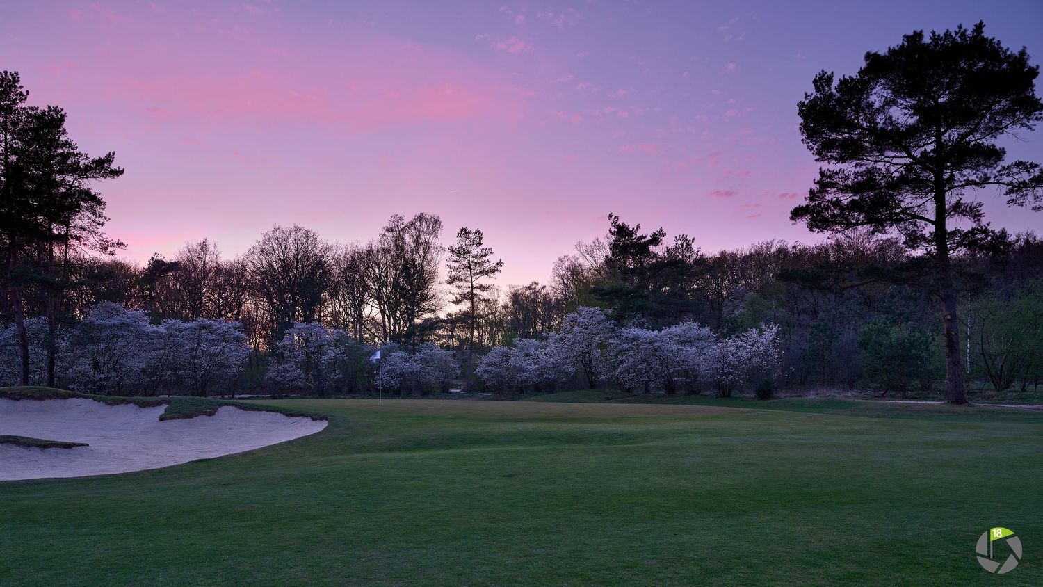 Golf course at dusk with sand bunker, lush green fairway, and blooming trees under a pink and purple twilight sky.