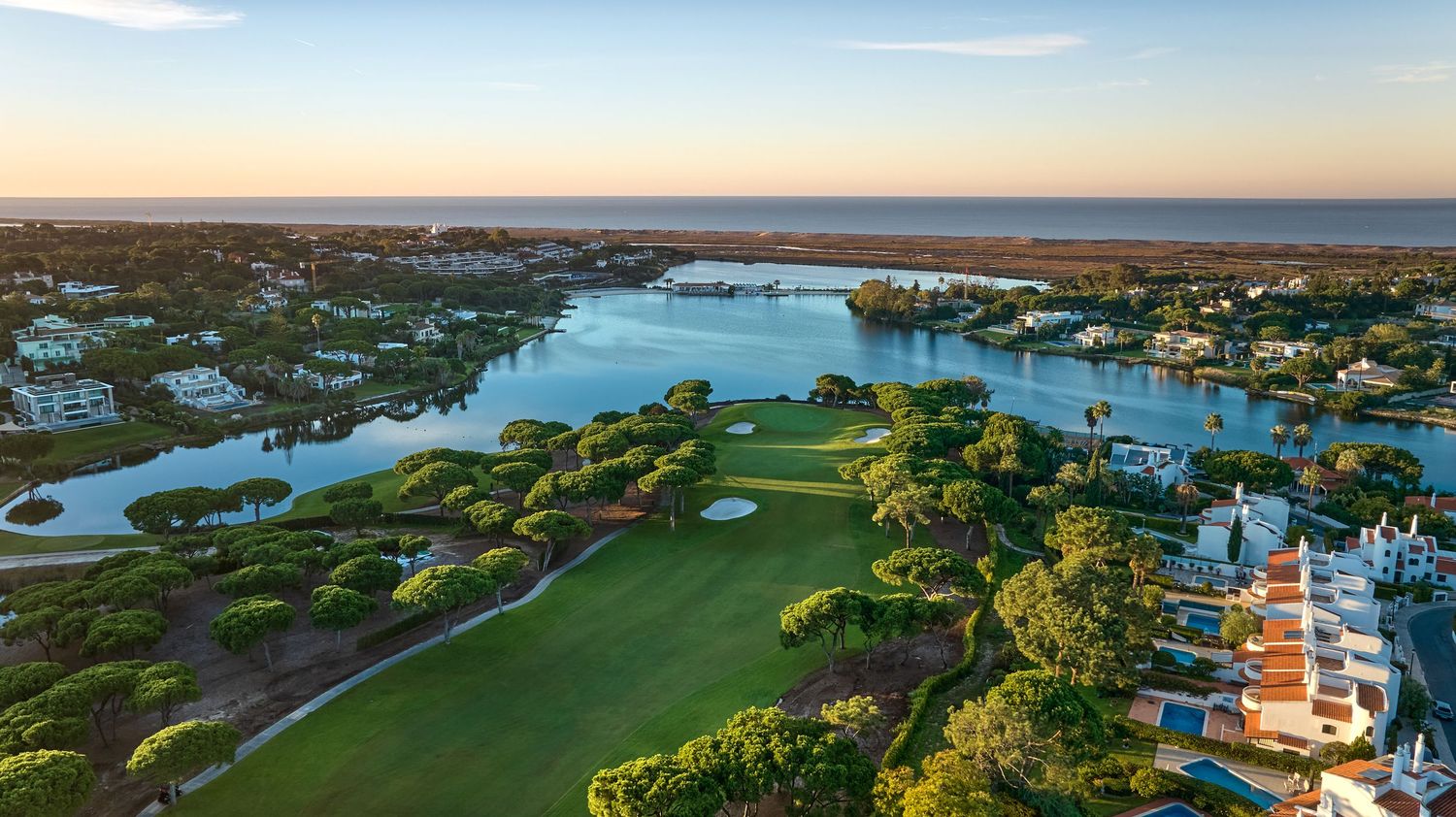 Aerial panoramic view of coastal golf courses and waterfront homes along the shoreline at sunset.