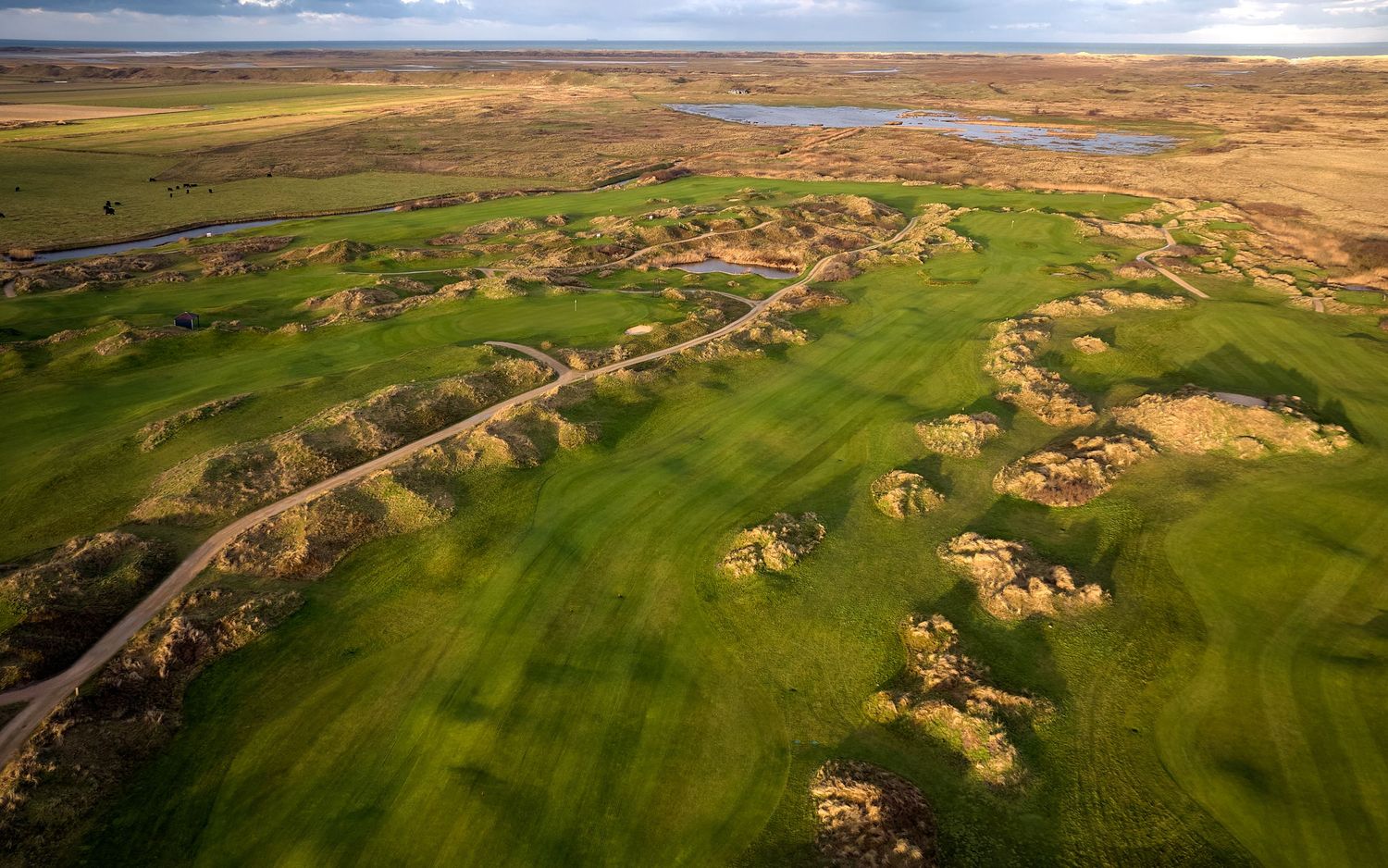 Aerial view of a lush green links golf course with natural rough terrain and a water hazard under dramatic skies.
