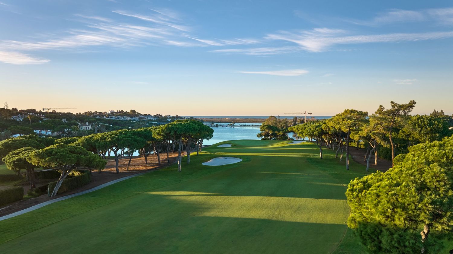 Aerial view of a lush golf course with manicured greens, sand bunkers, and pine trees under a clear blue sky.