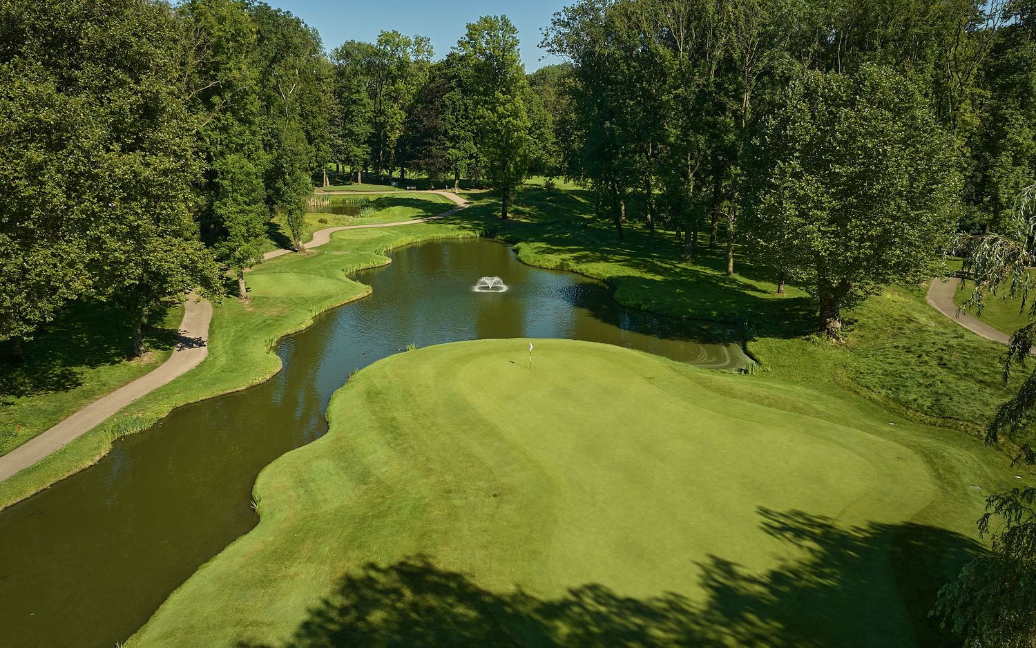 Aerial view of a lush golf course fairway with a winding pond, manicured greens, and tree-lined borders on a sunny day.