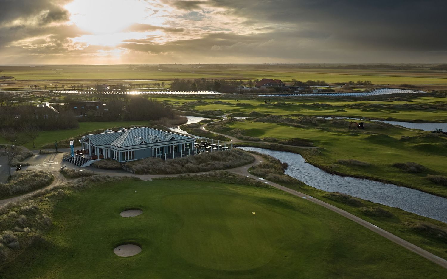 Aerial view of a golf course clubhouse at sunset with winding water features and lush greens stretching to the horizon.