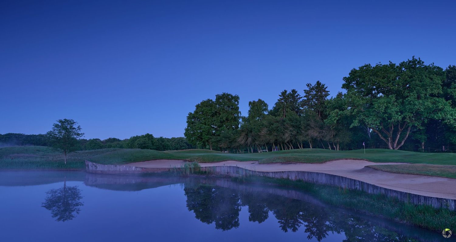 A tranquil golf course at dusk with a reflective water hazard and tree-lined fairway under deep blue skies.