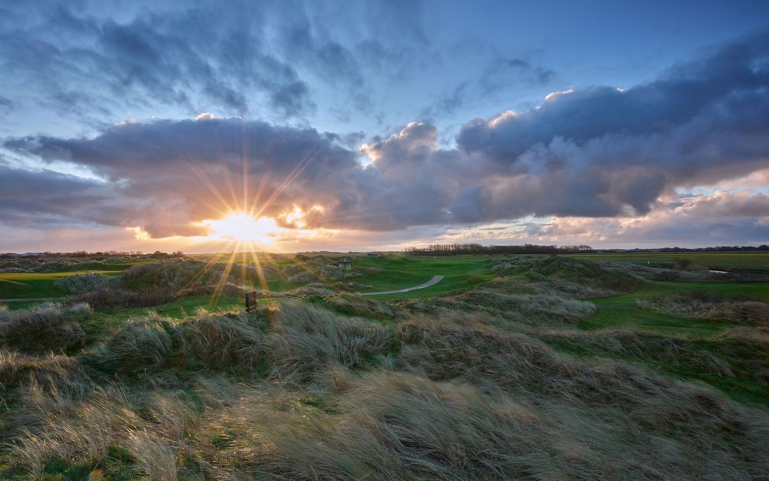 Sun bursting through dramatic clouds over a lush links golf course with windswept dunes at sunset.