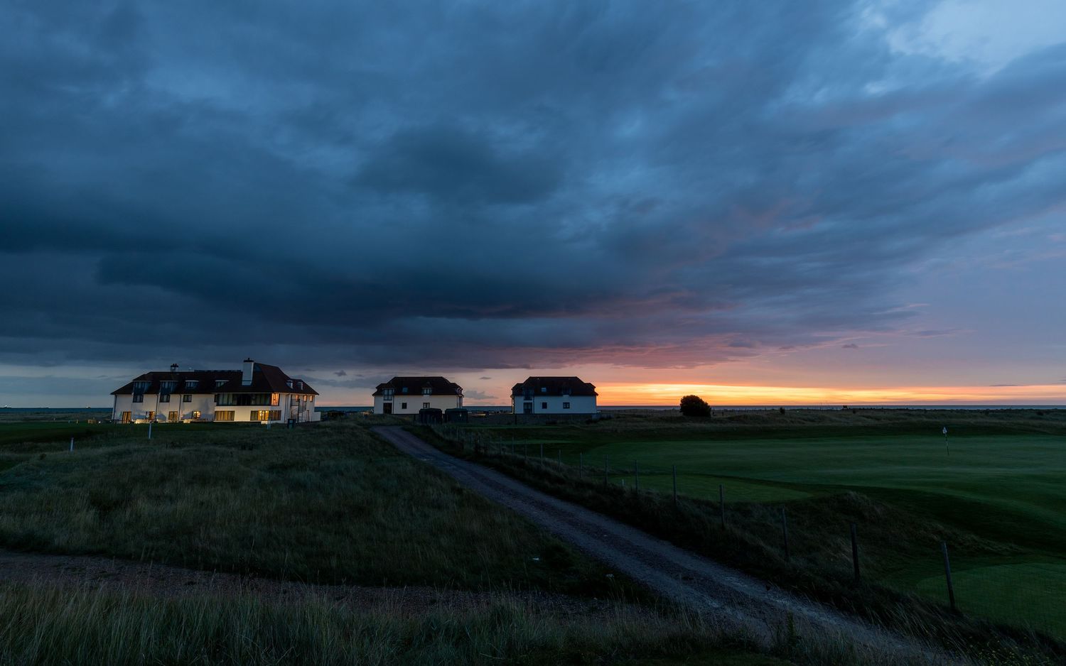 Rural farmhouses at dusk under dramatic storm clouds with a vivid sunset glowing on the horizon over green fields.