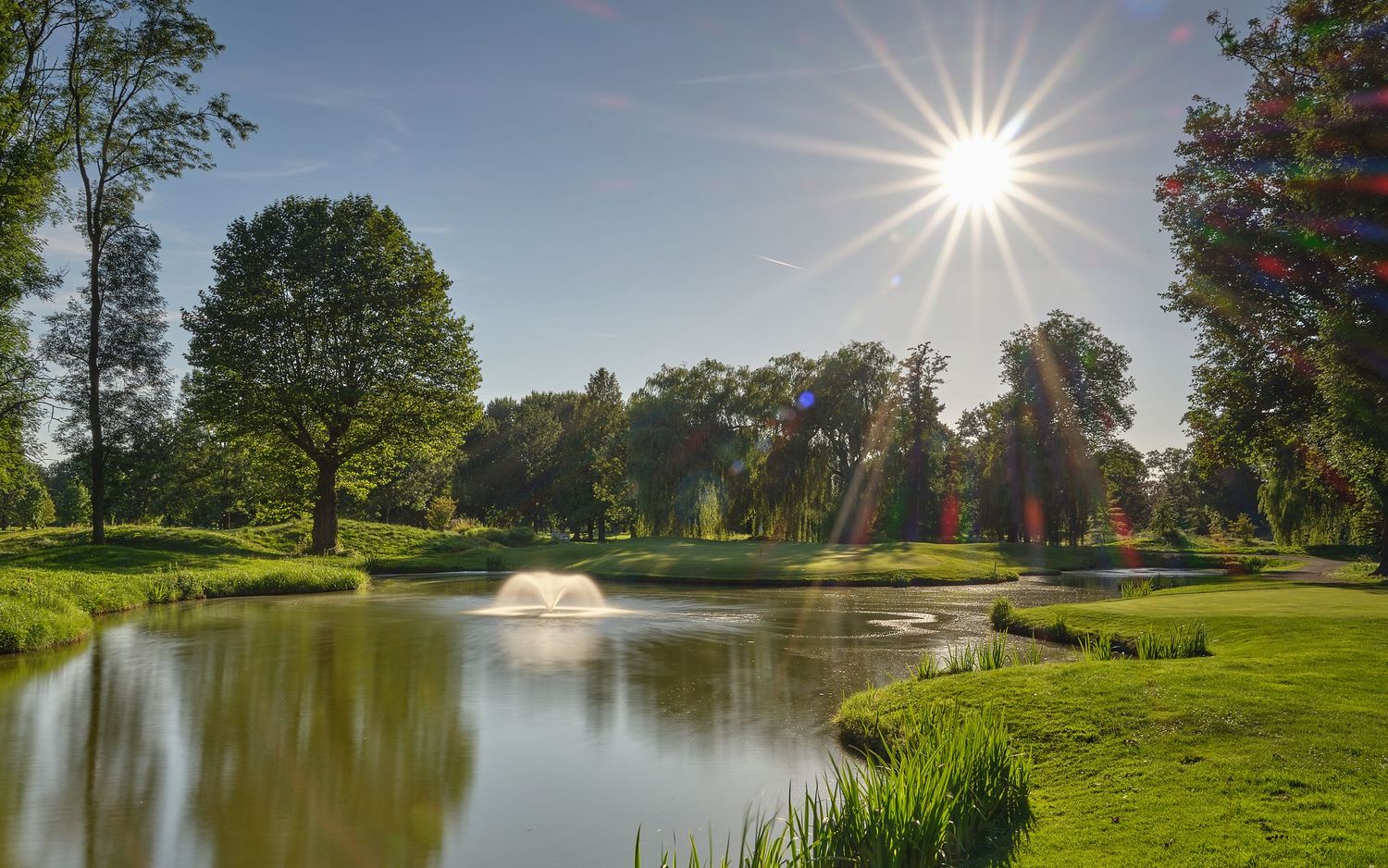 Sunny golf course pond with fountain, lush green trees, and bright sunburst sky reflected in calm water.