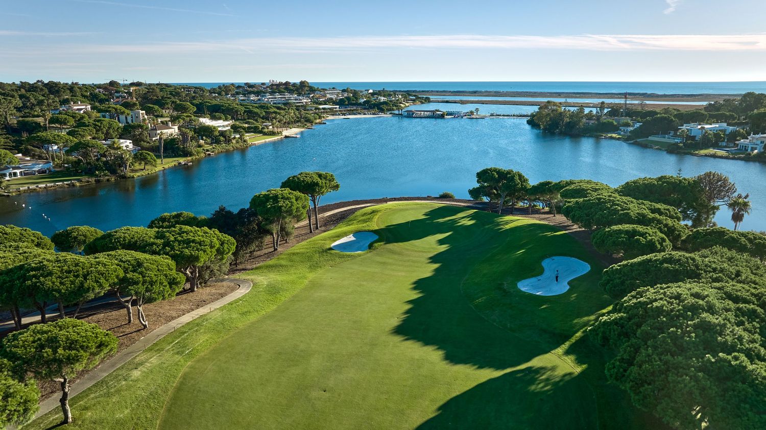Aerial view of a lush golf course with sand bunkers beside a serene blue lagoon surrounded by pine trees.