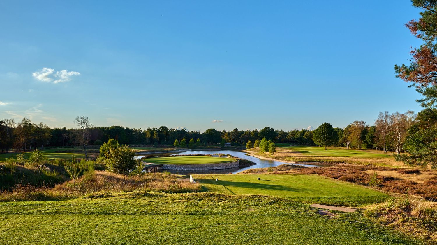 Scenic golf course fairway with water hazard, lush greens, and tree-lined landscape under a clear blue sky.