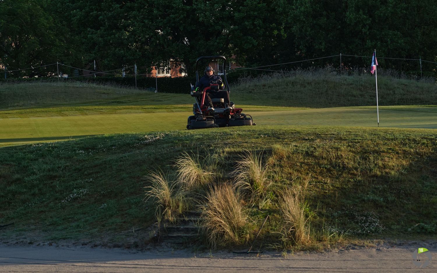 Groundskeeper mowing grass on a golf course at dusk near a sand trap and flag pin.