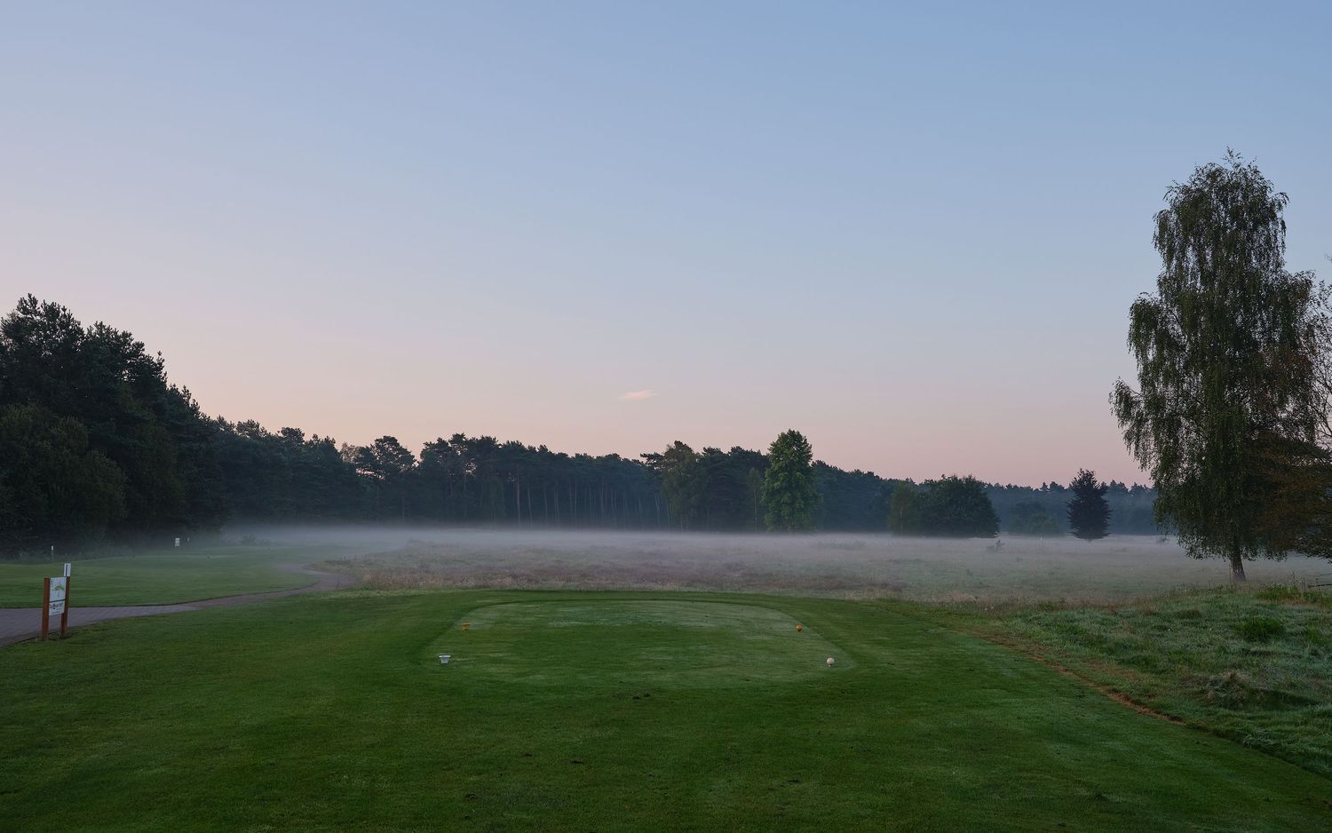 Misty morning sunrise over a green golf course fairway with trees silhouetted against the dawn sky.