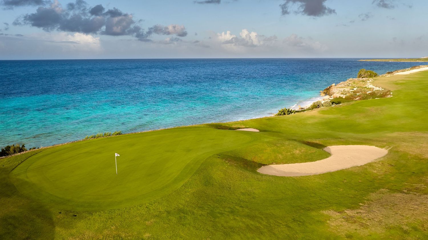 A panoramic view of a coastal golf course with emerald greens, white sand bunkers and turquoise ocean waters.