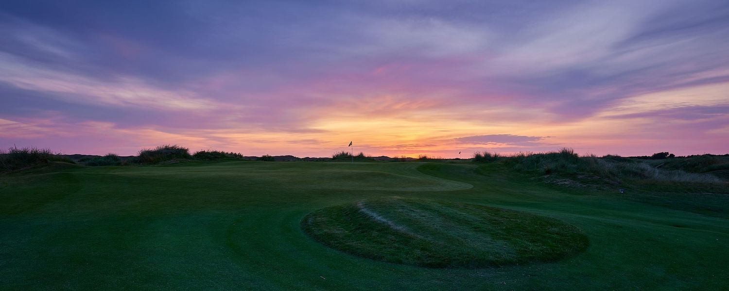 Golf course at sunset with dramatic purple and orange sky illuminating the green fairway and sand bunkers.