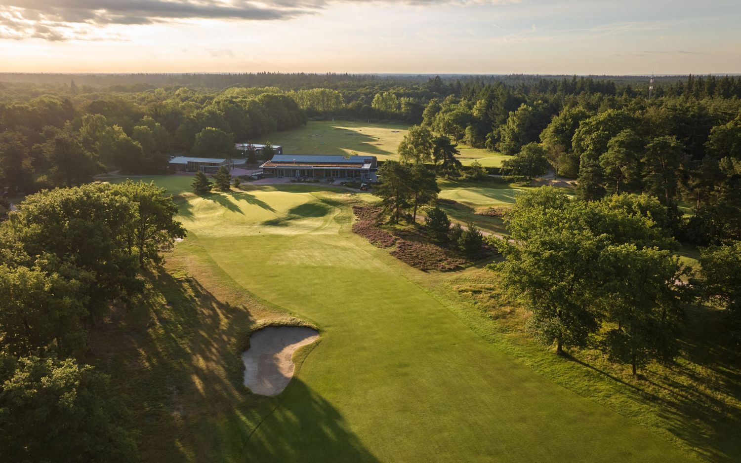 Aerial view of a lush golf course at sunset, featuring manicured fairways, sand bunkers, and a clubhouse surrounded by trees.