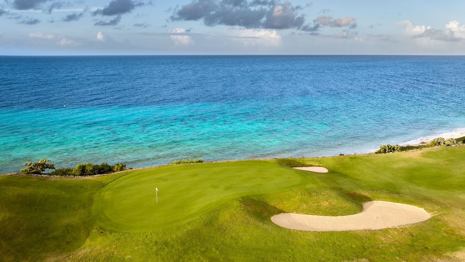 A panoramic view of a coastal golf course with rolling green fairways, sandy bunkers and turquoise ocean waters.