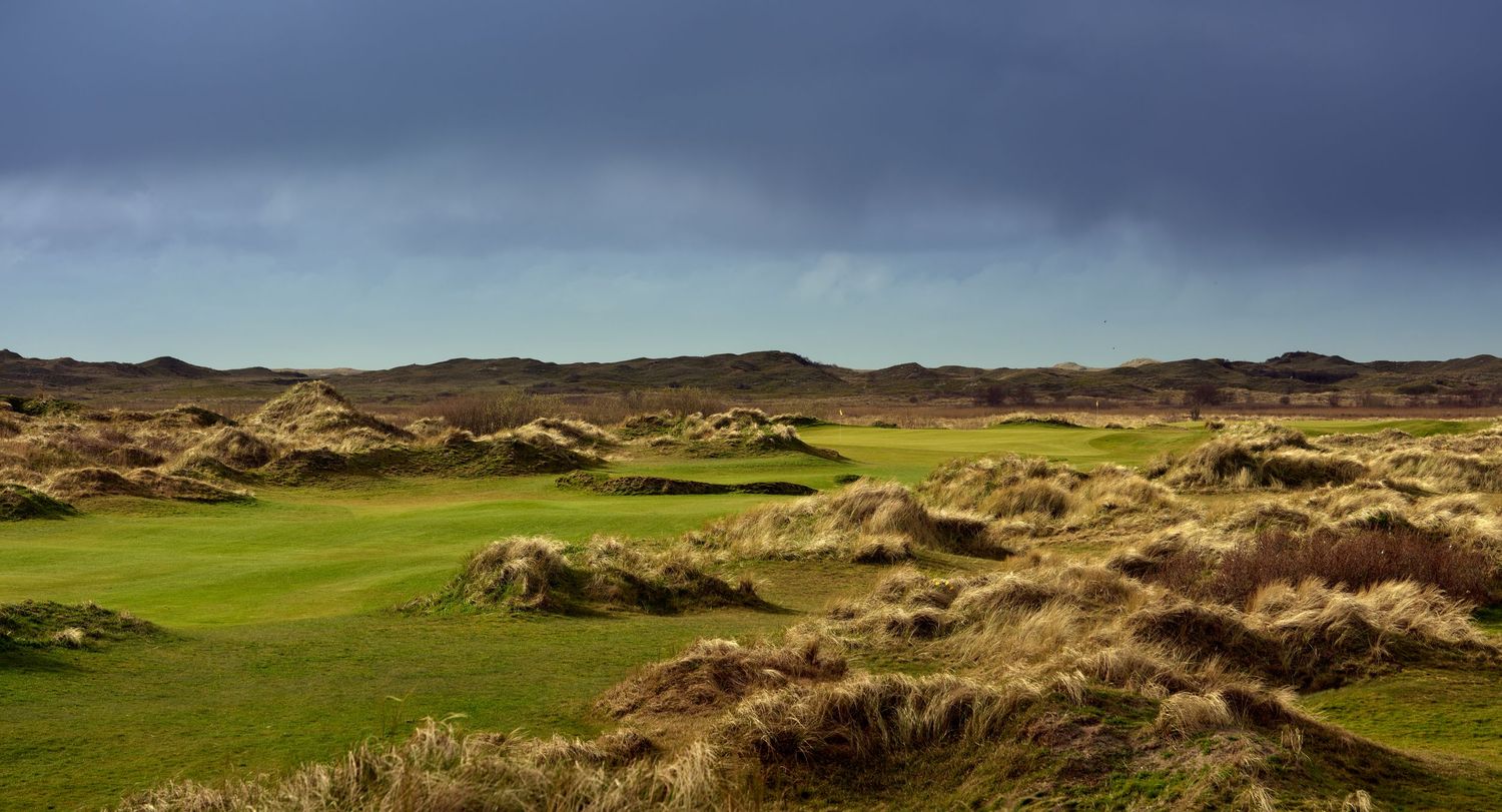 Links golf course with lush green fairways surrounded by sandy dunes under dramatic stormy skies.
