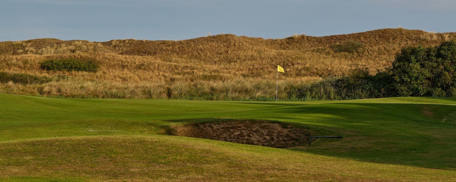 Golf course fairway with yellow flag, sand bunker, and golden dunes under clear blue sky.