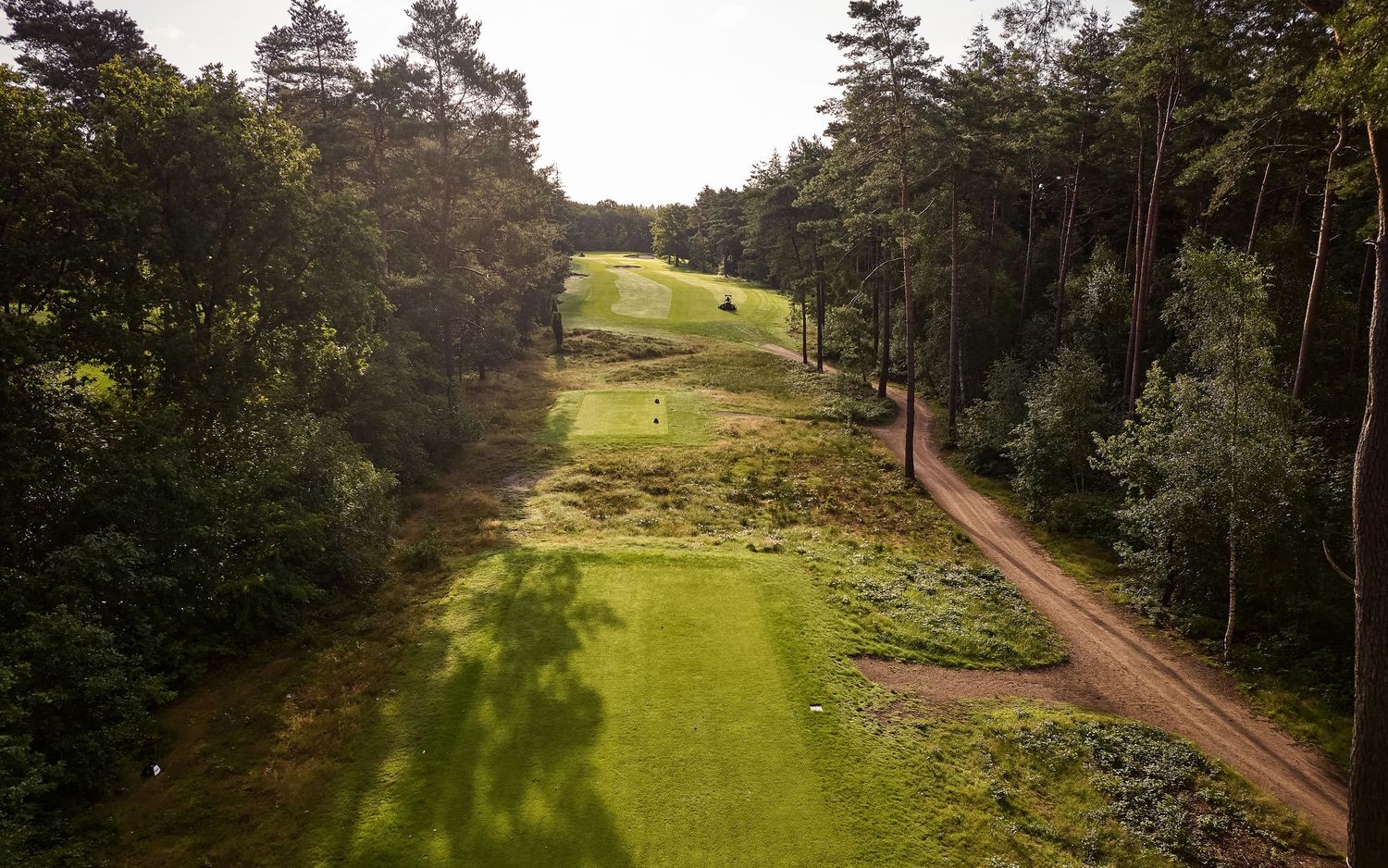 Aerial view of a lush green golf course fairway surrounded by dense forest trees and a dirt path on a cloudy day.