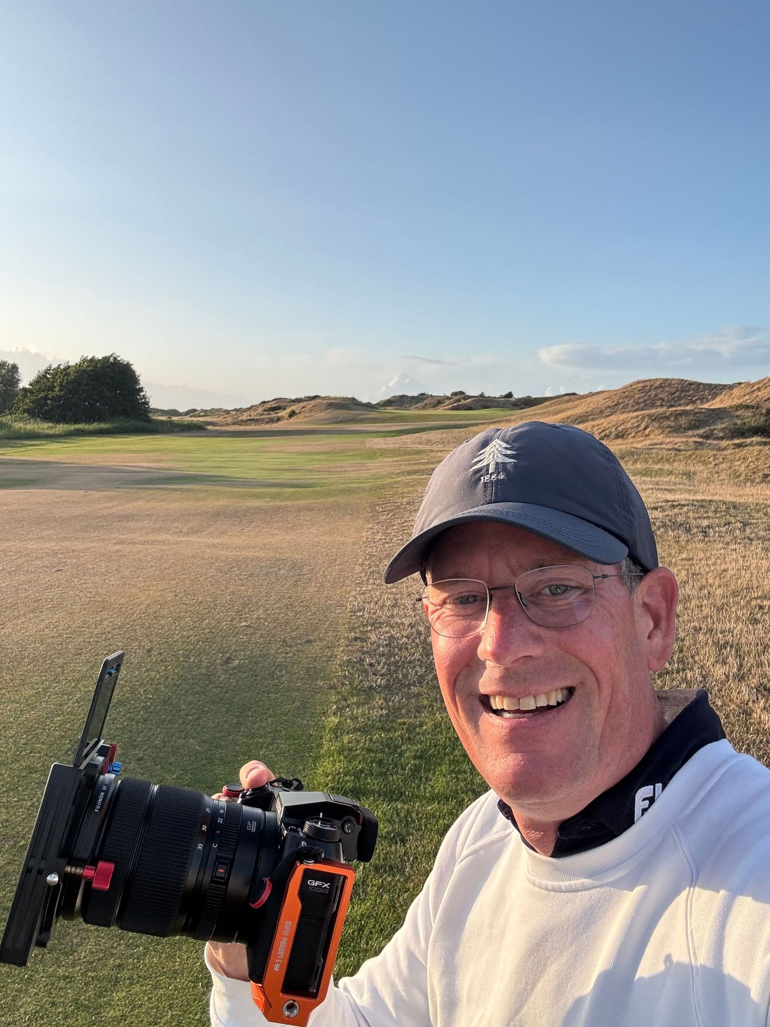 Smiling man holding a professional camera on a sunny golf course with rolling dunes and fairway in the background.