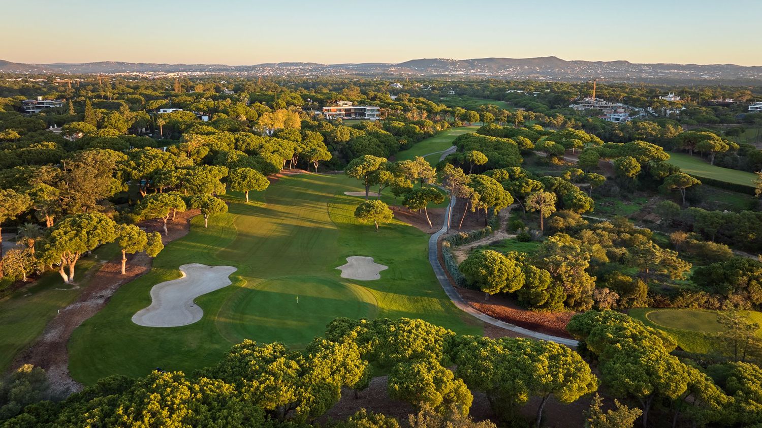 Aerial view of a lush golf course with sand bunkers and winding paths surrounded by green trees at sunset.