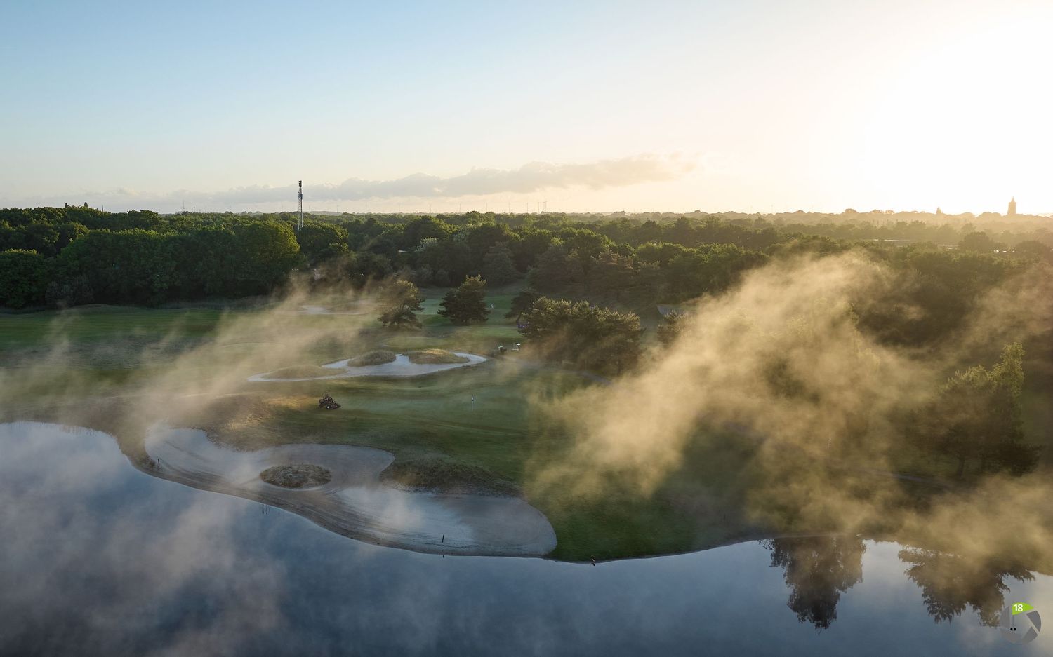 Aerial view of a misty golf course at sunrise with fog drifting over lush green fairways and a serene water hazard.