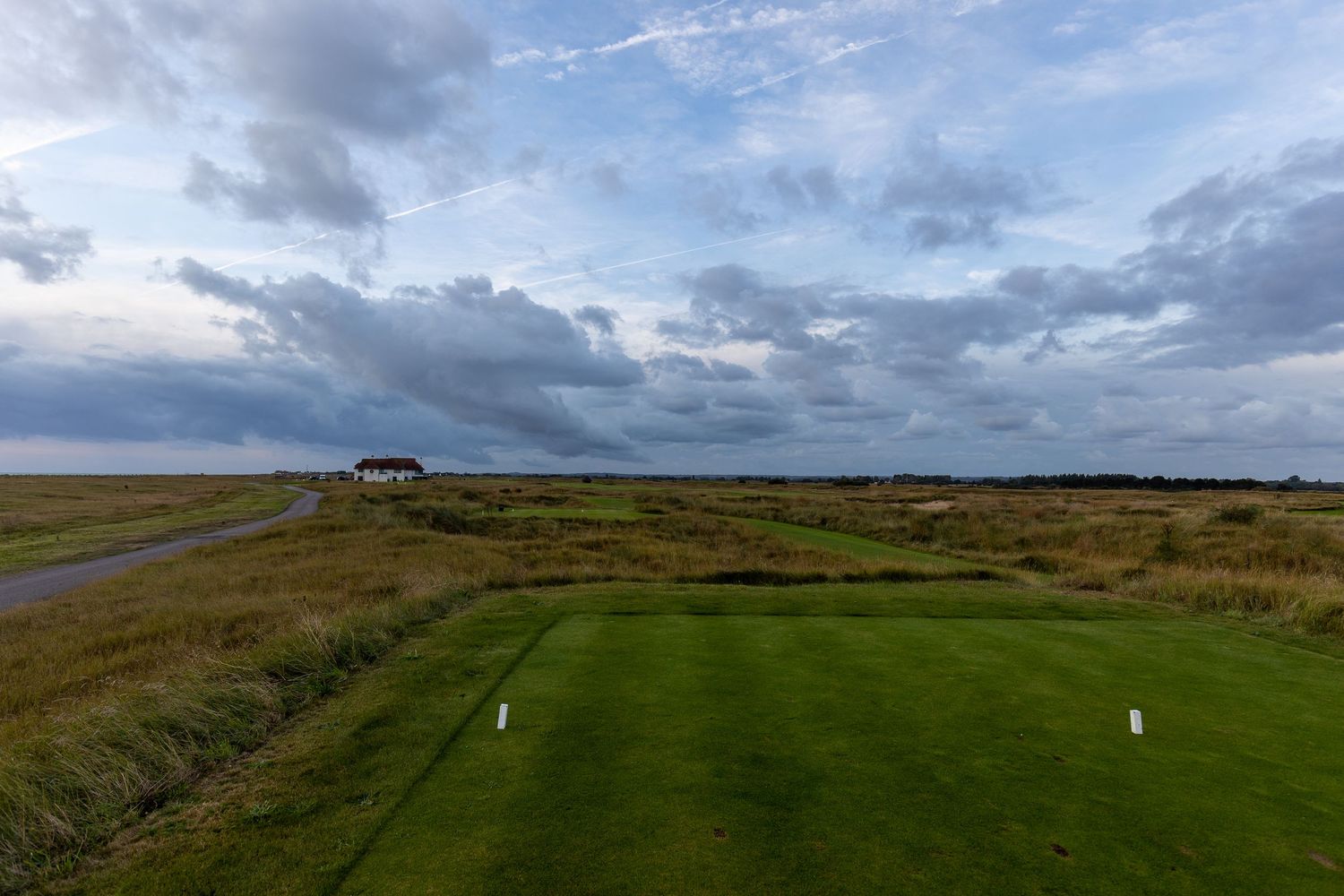 Aerial view of a links golf course with lush green fairways, wild rough grass, and a cloudy sky over a coastal landscape.