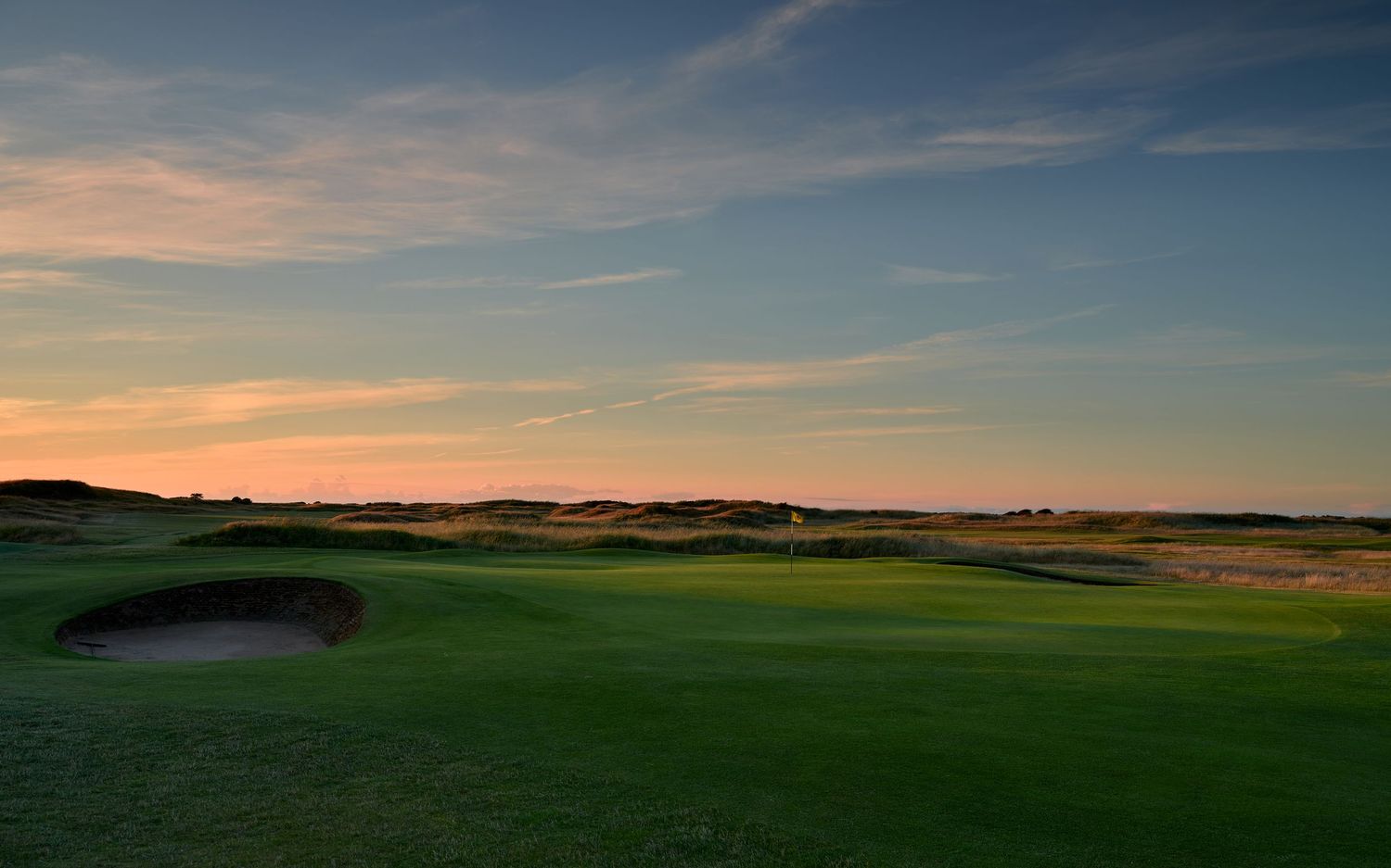 Scenic golf course at sunset with lush green fairways, sand bunkers, and a colorful sky over a links-style course.