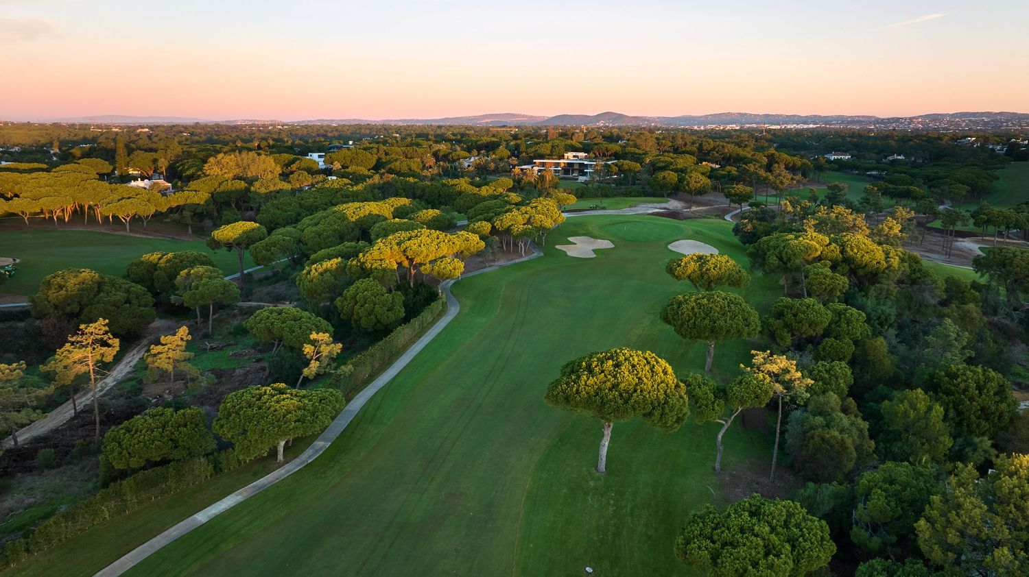Aerial view of a scenic golf course surrounded by lush trees at golden sunset with mountains in background.