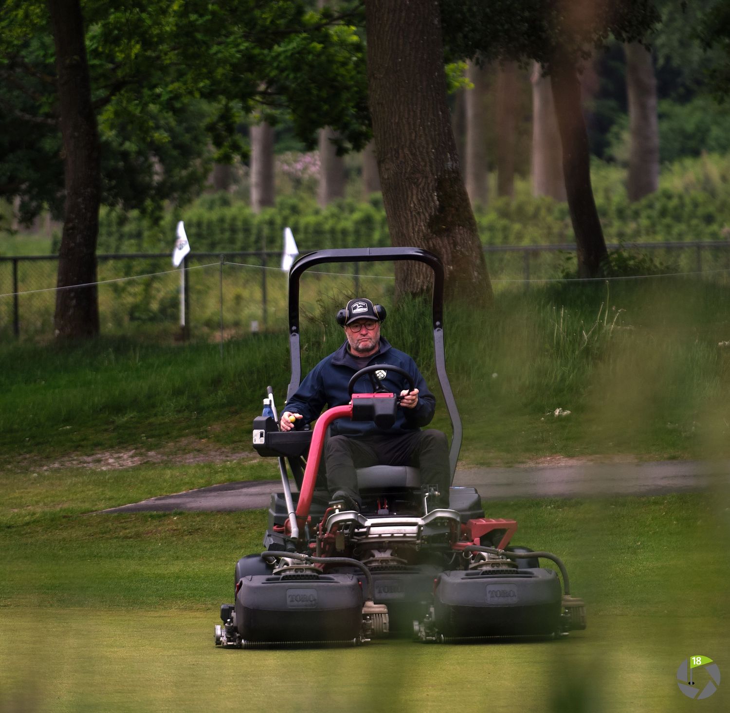 Groundskeeper riding a large commercial lawn mower on a lush green golf course surrounded by trees.
