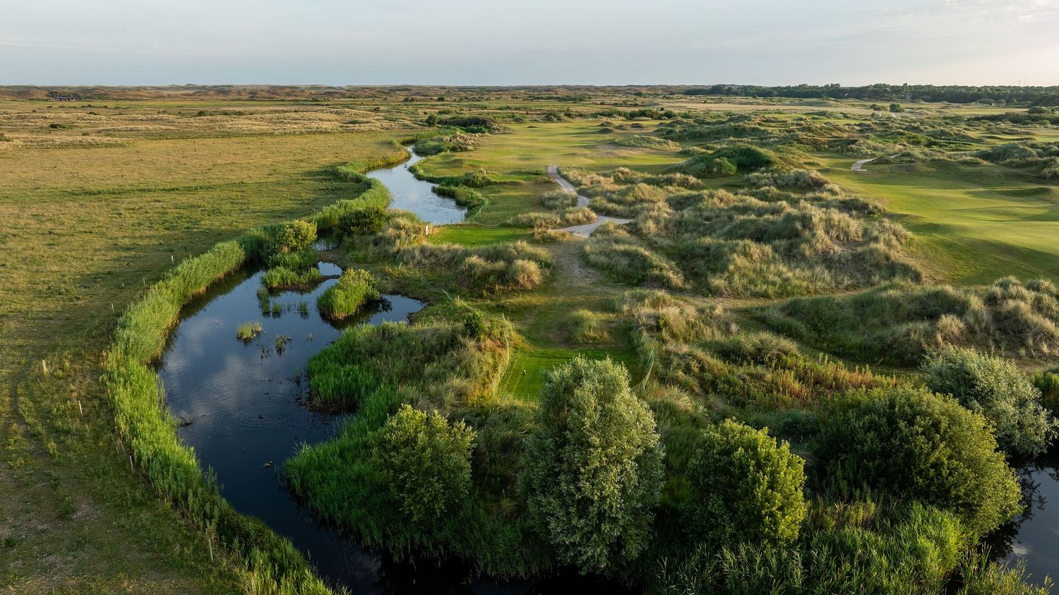 Aerial view of a scenic links golf course with winding river, lush green fairways, and natural wetlands under soft evening light.