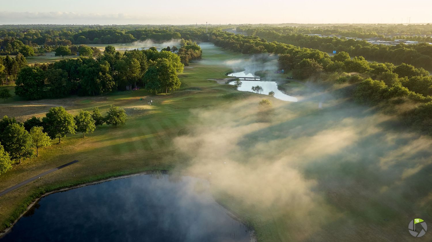 Aerial view of a misty golf course at sunrise with ponds, fairways, and trees stretching to the horizon.