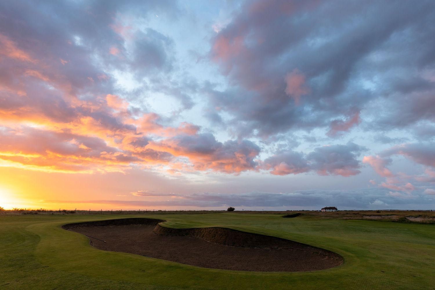 Stunning sunset over a links golf course with sand bunkers, dramatic orange and blue cloudy sky, and coastal views.