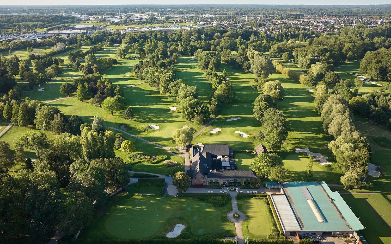 Aerial view of a lush green golf course with manicured fairways, clubhouse, and surrounding trees near a town.