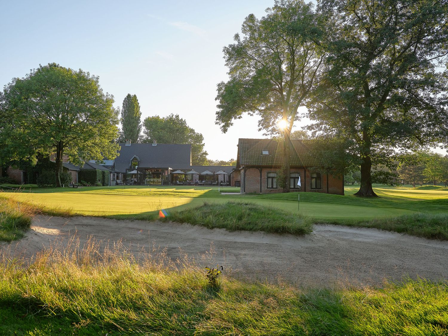 Sunlit golf course with sand bunker, lush green fairway, and rustic clubhouse buildings surrounded by tall trees.
