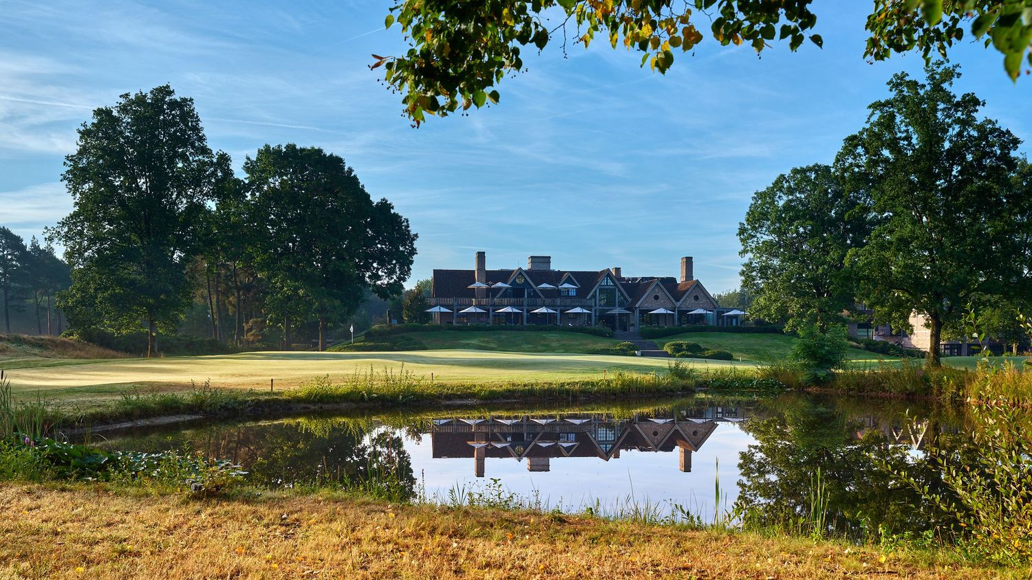 Elegant brick clubhouse reflected in a tranquil pond surrounded by lush trees on a sunny golf course.