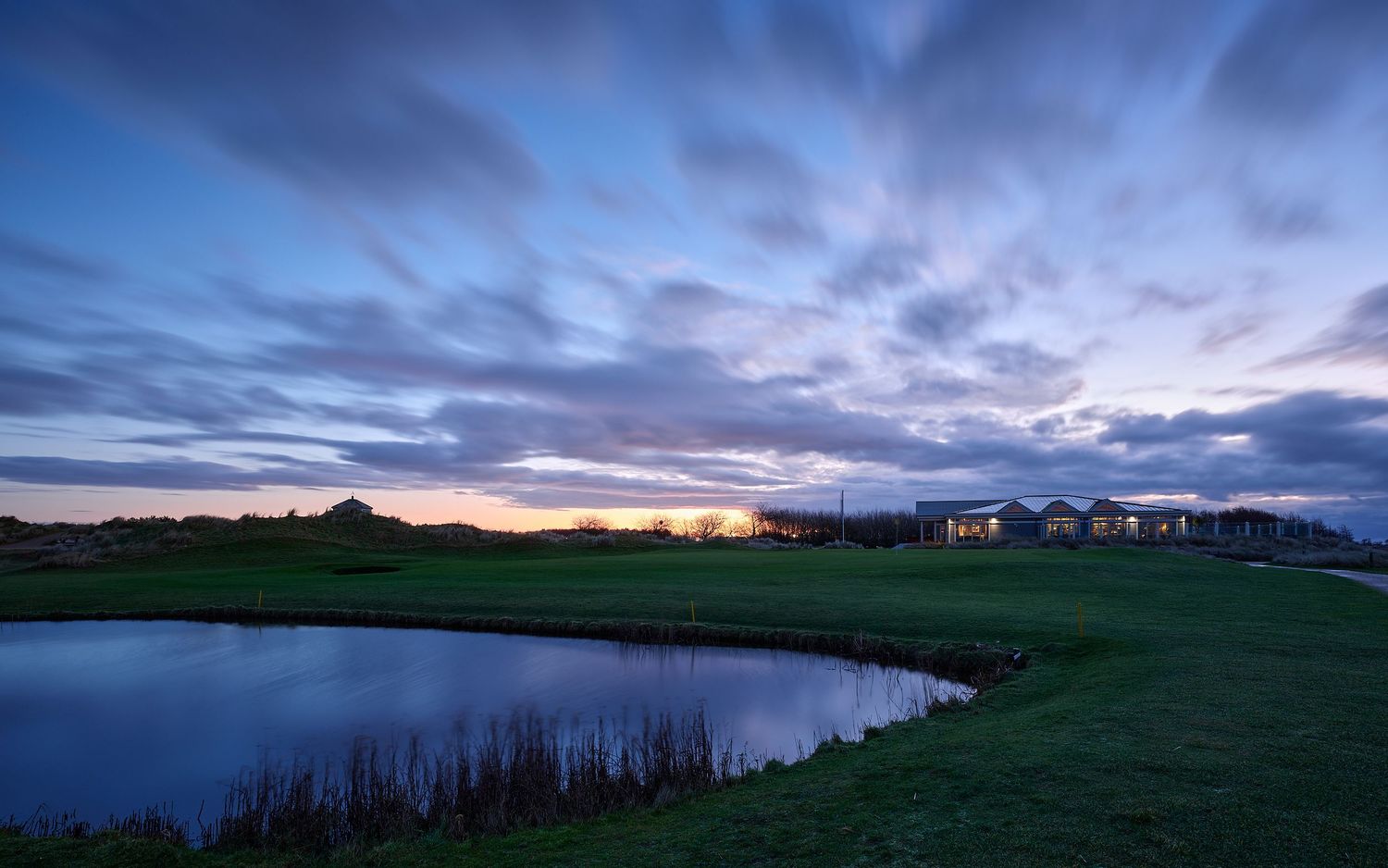 Twilight golf course with serene pond, lush green fairways, and dramatic cloudy sky at sunset.