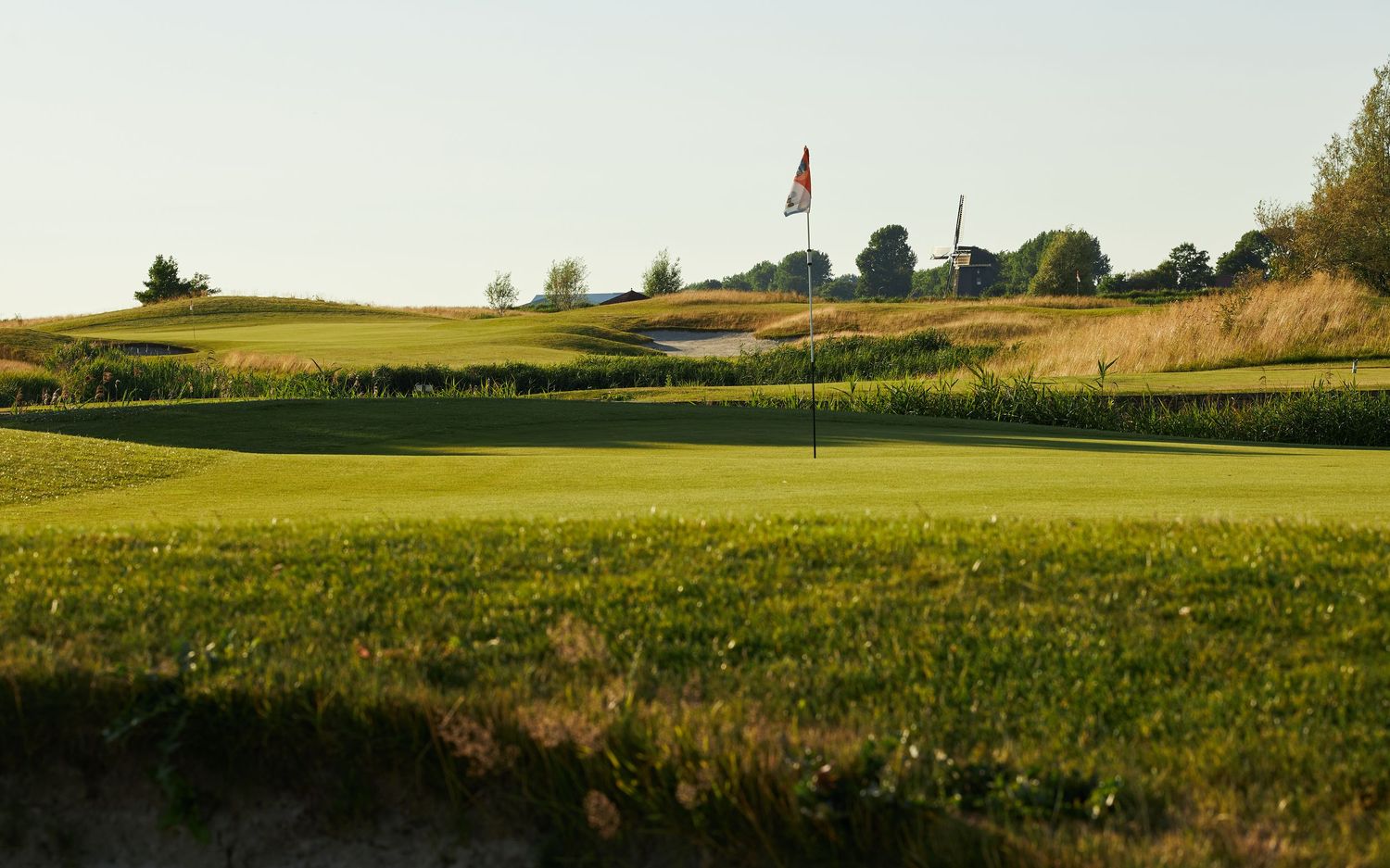 Rolling green hills stretch across a scenic rural landscape with a distant farm and flag pole visible on the horizon.