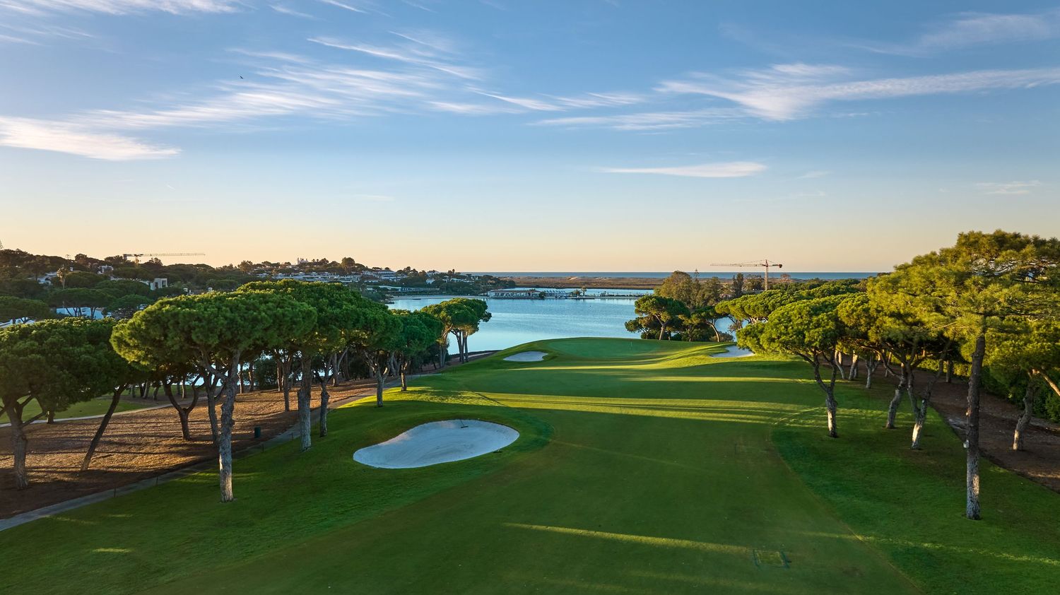 Aerial view of a lush golf course fairway with sand bunkers, pine trees, and a scenic lake under a clear blue sky.