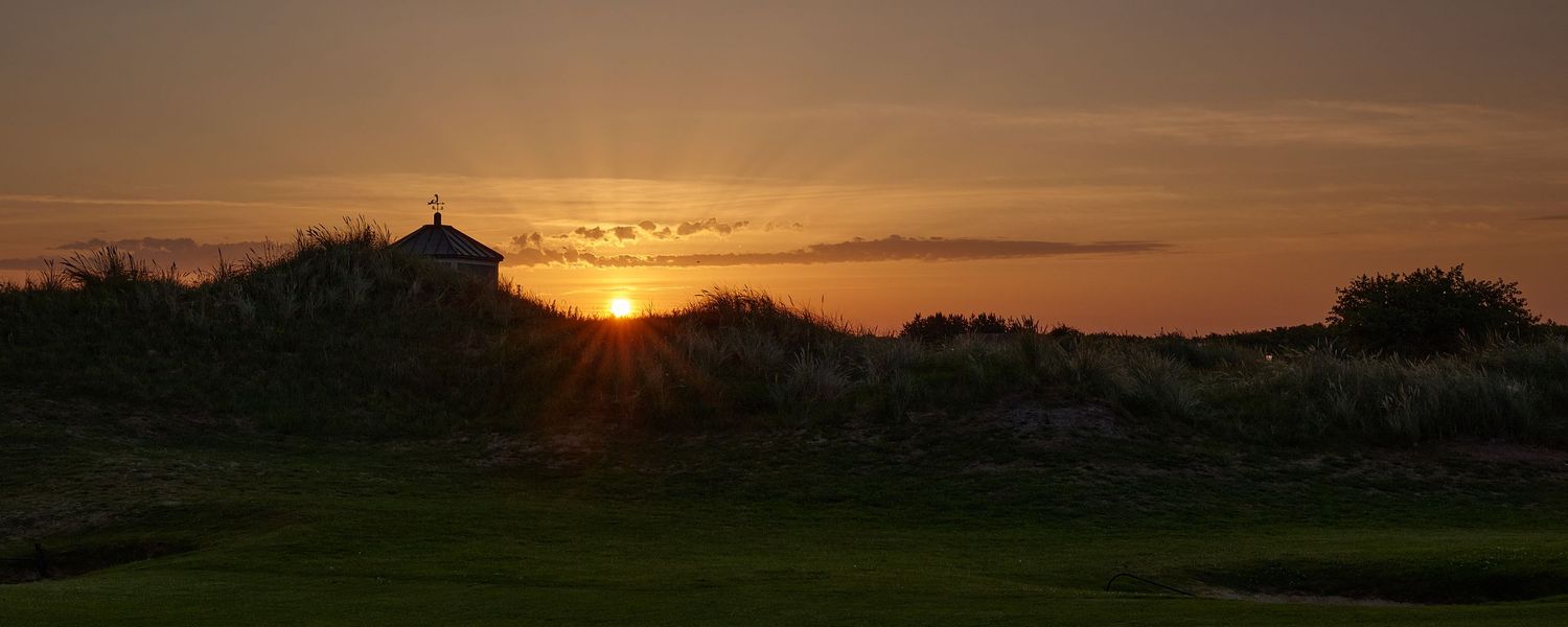 Golden sunset silhouettes a small chapel amid coastal dunes on a tranquil golf course.
