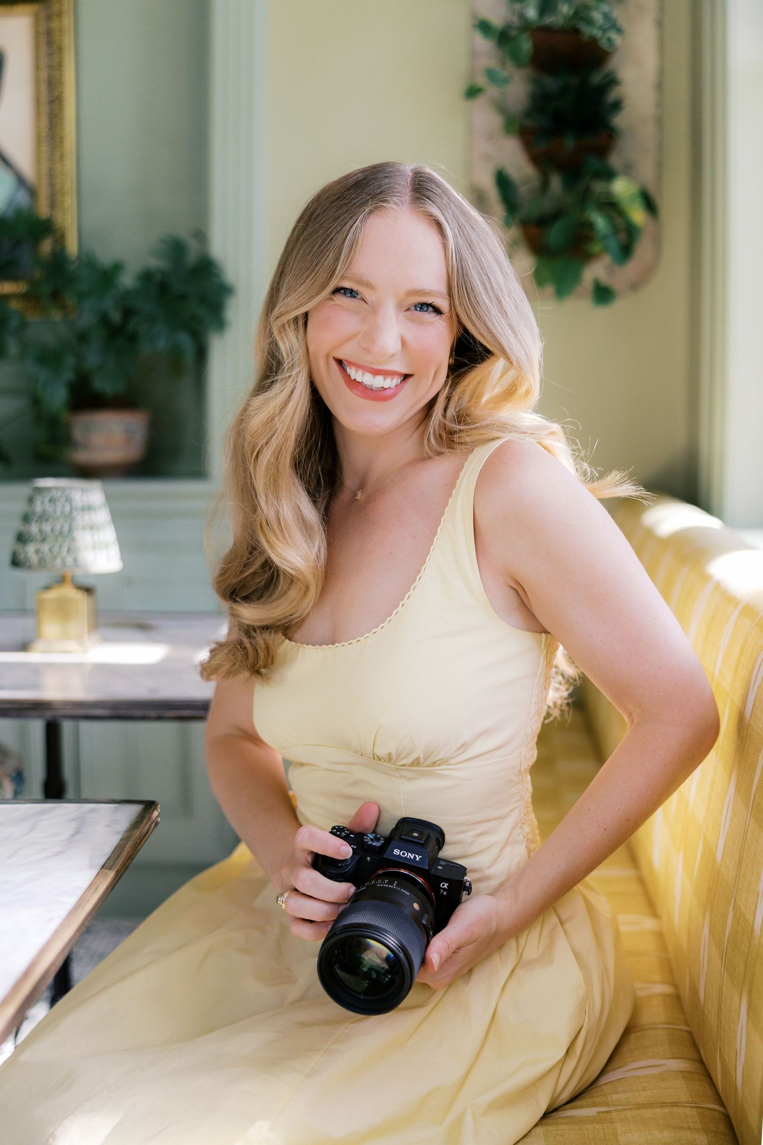 Smiling blonde woman in yellow dress holding a DSLR camera, seated on a yellow striped sofa in elegant room.