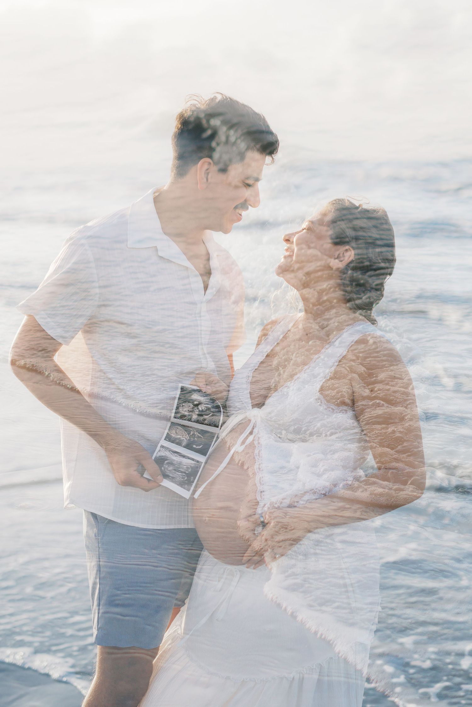 Couple sharing a tender moment on the beach, holding ultrasound photos, in a dreamy double exposure maternity portrait.