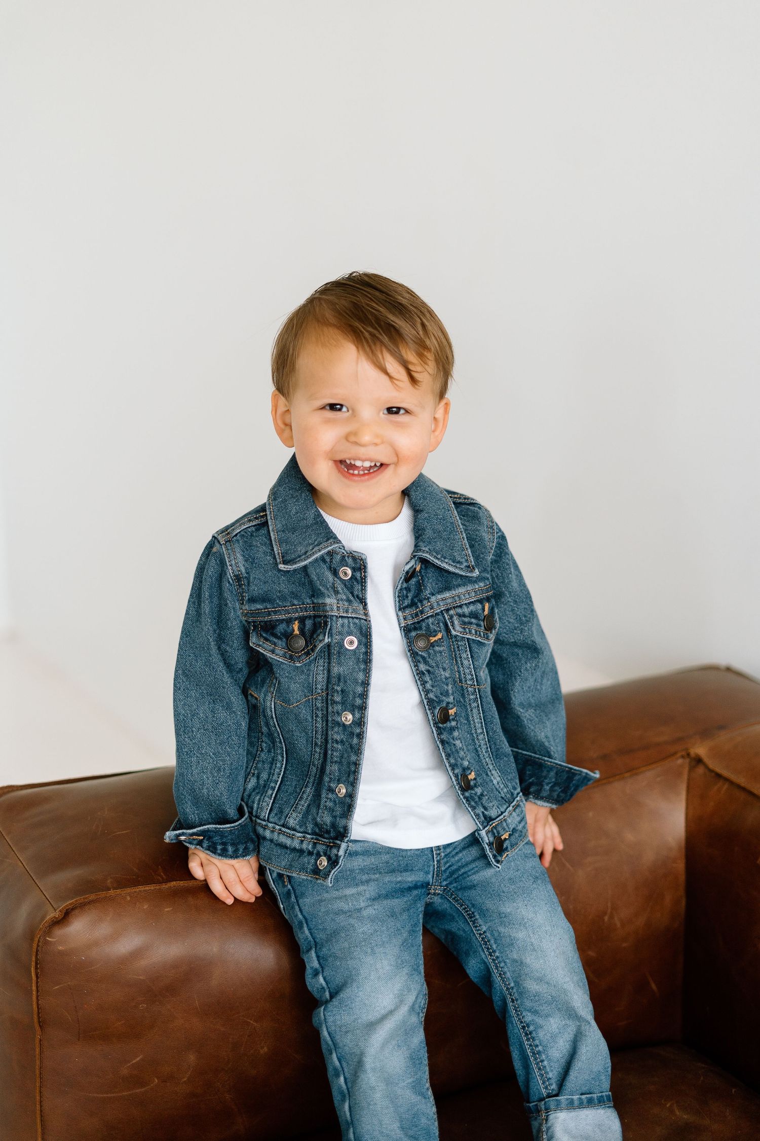 Child wearing denim jacket and jeans smiling on brown leather couch.