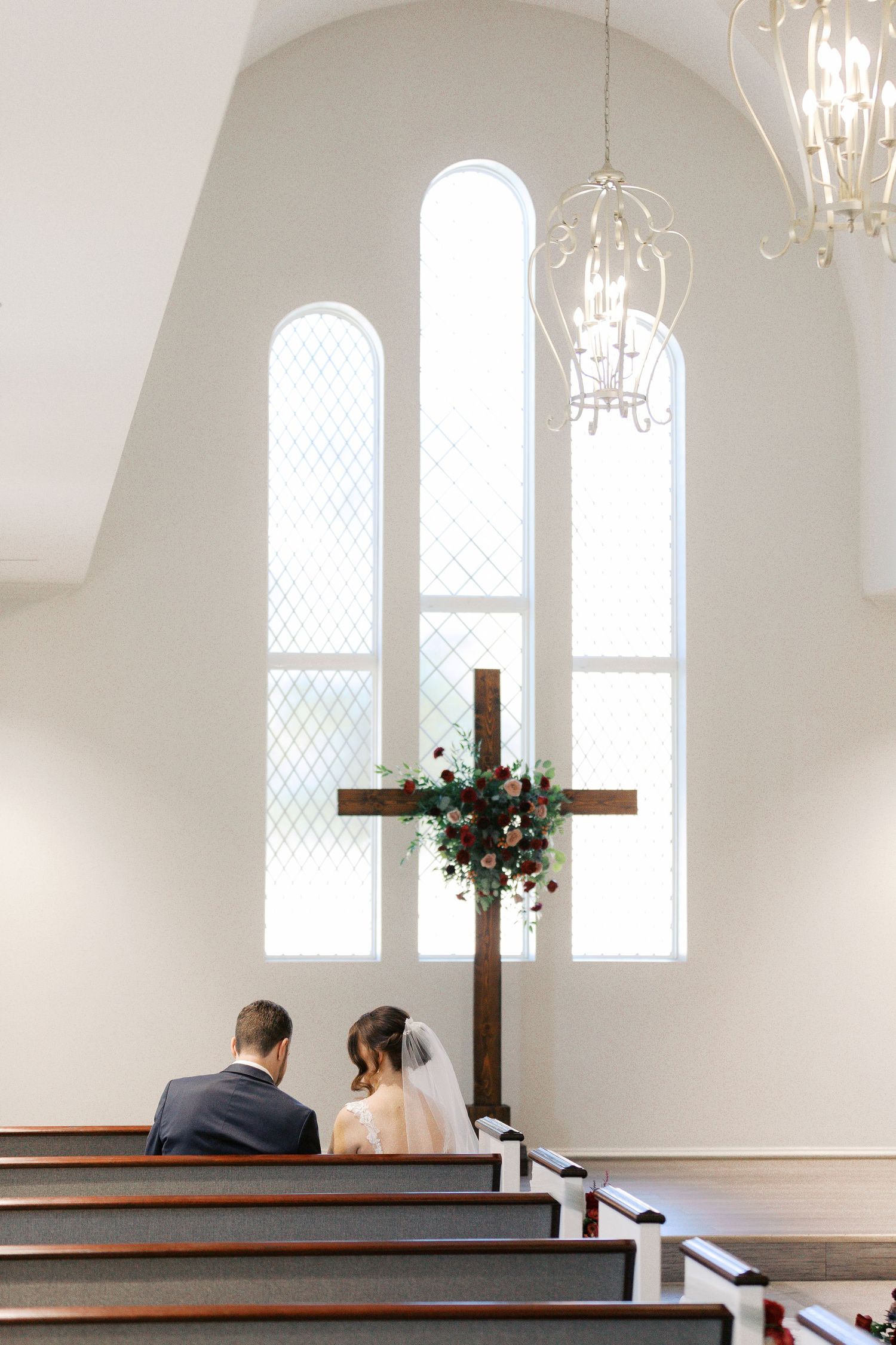 A couple sits together in an elegant white church with arched windows and chandeliers.