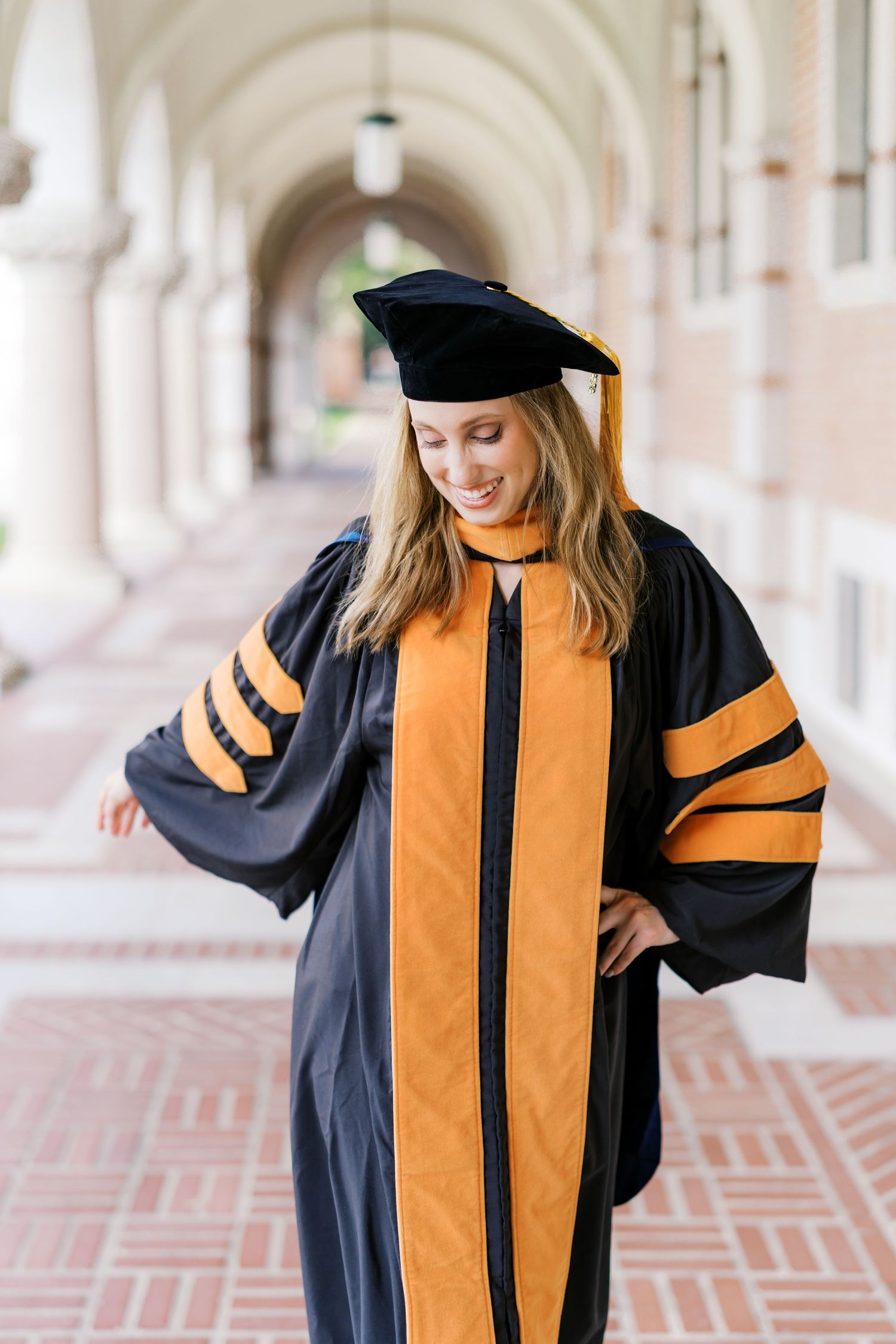 Graduate in doctoral robes with orange accents poses confidently in university corridor.