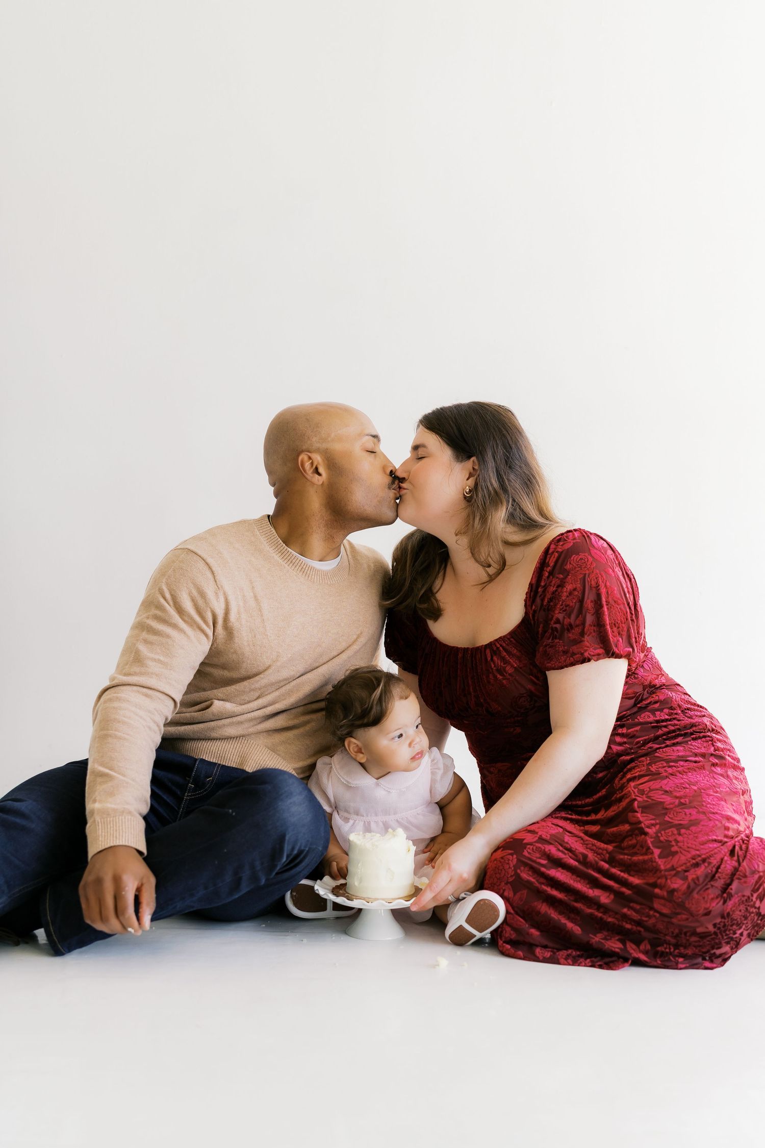 A couple in casual attire shares an intimate moment with their baby on a white background.