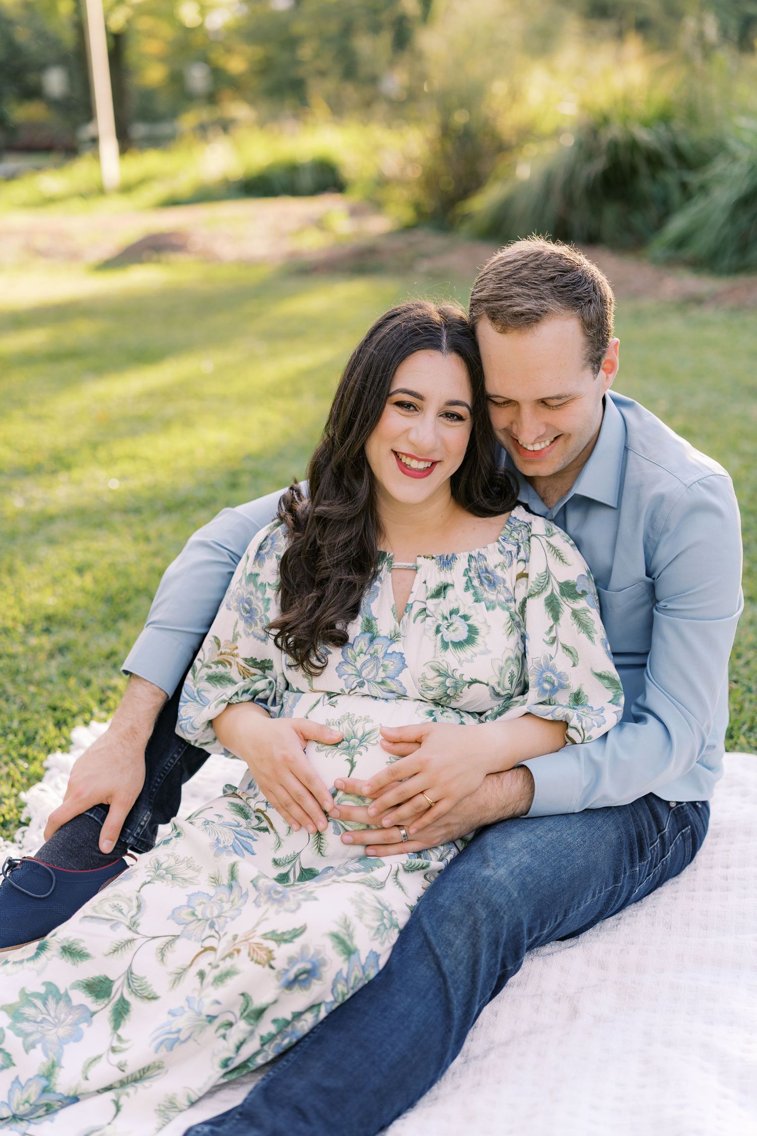 Couple relaxing on white blanket in park during sunset maternity photo session.